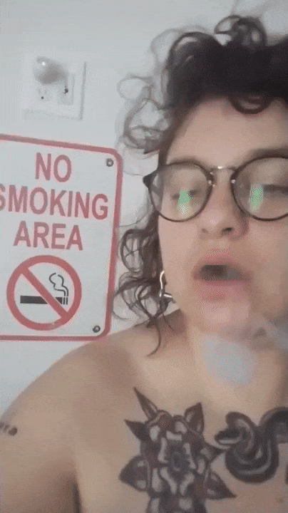 gif of me blowing smoke at the camera, i am white with wavy brunette hair, wearing dark glasses, and have visible tattoos, there is a sign behind me that says: NO SMOKING